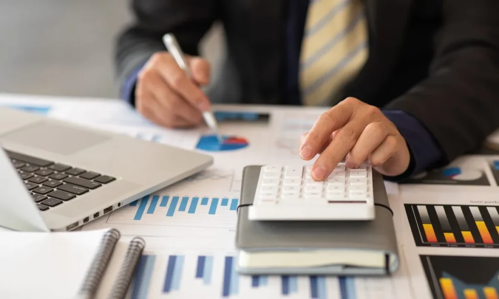 A close-up of a person in a business suit using a white calculator and a pen to analyze financial charts and graphs on a desk. A laptop and several notebooks are also visible, suggesting a detailed budget or data review.