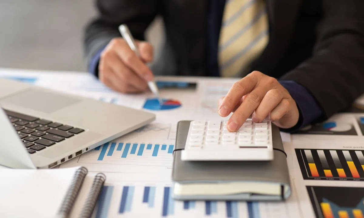 A close-up of a person in a business suit using a white calculator and a pen to analyze financial charts and graphs on a desk. A laptop and several notebooks are also visible, suggesting a detailed budget or data review.