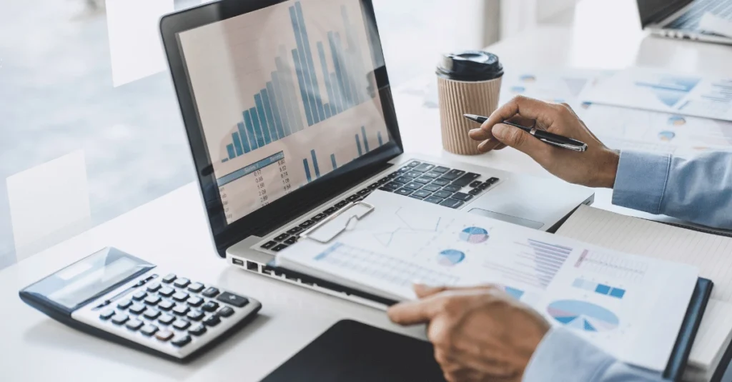 An office professional analyzing data on a laptop and printed reports featuring various charts and graphs, with a calculator and coffee on the desk.